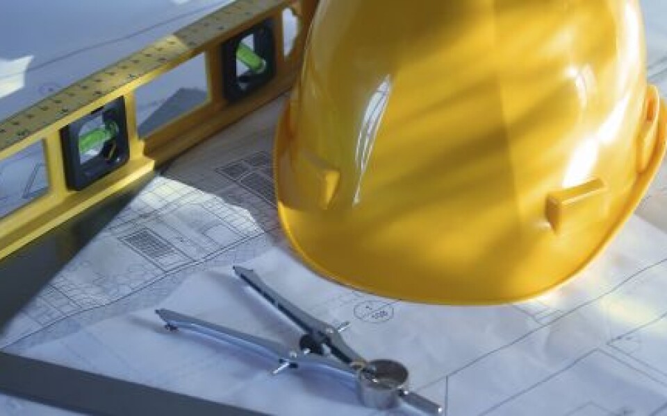 Casque de chantier jaune posé sur des plans d'architecte, avec un niveau à bulle et un compas